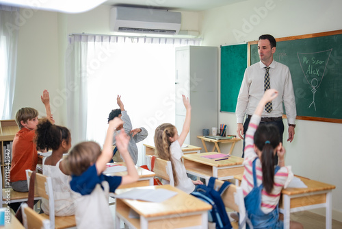 Elementary school students raising hands during classroom participation while teacher observes in bright educational environment with collaborative learning atmosphere.