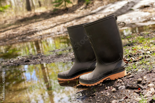 Tableau sur toile Pair of black rubber boots standing near the muddy puddle in the bushy backyard in spring wet weather