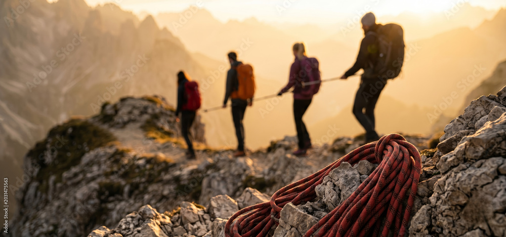 Fototapeta premium Four adult mountaineers traverse a rocky alpine trail at golden hour, connected by a safety rope, with coiled climbing gear resting on nearby rocks.