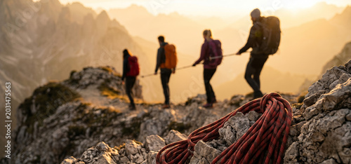 Four adult mountaineers traverse a rocky alpine trail at golden hour, connected by a safety rope, with coiled climbing gear resting on nearby rocks.