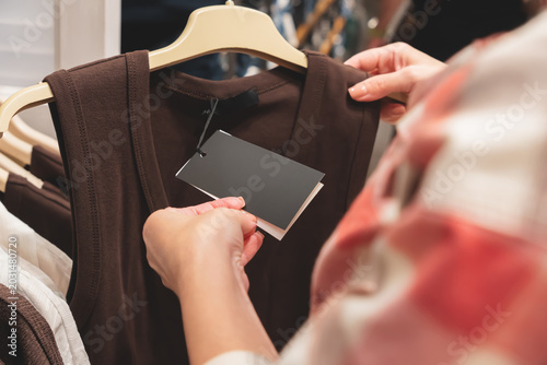 A girl in a shopping mall holds a hanger with a brown top in one hand, and holds a tag with the other hand.Clothing item hangs in store setting. Customer examines apparel in retail environment
