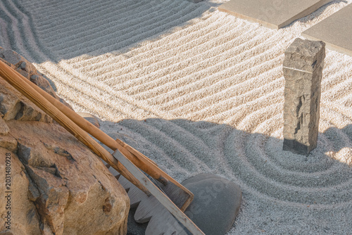 A peaceful zen garden with sand patterns and a stone pillar. Nearby there is a wooden rake for creating lines. Sunlight, meditative space.Зhoto is suitable for websites or print media.