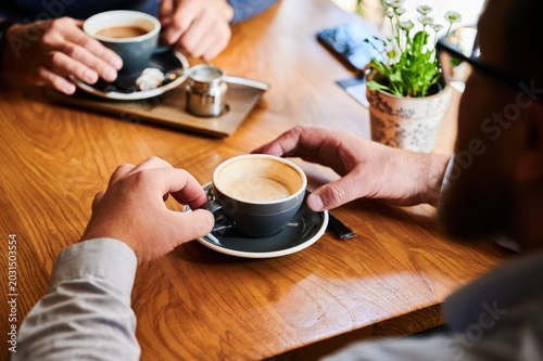 Man holding cappuccino at cafe table while another person prepares coffee in background, relaxed meeting scene with warm light, wooden surface and green plants