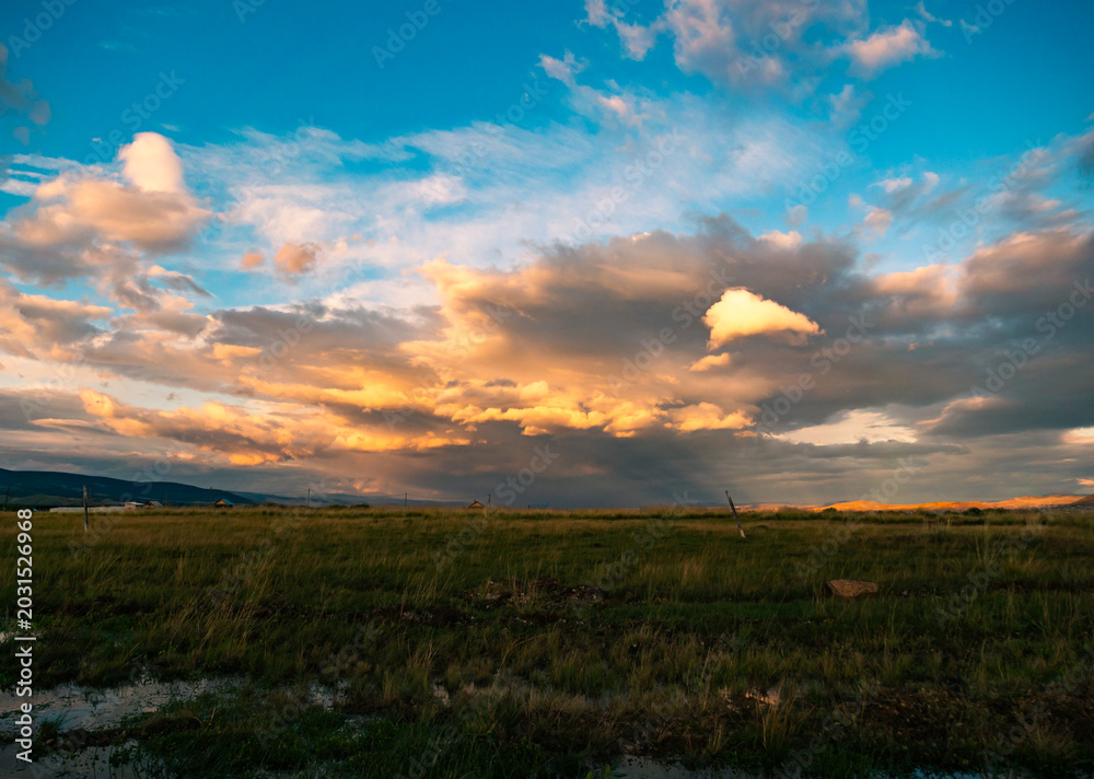 Obraz premium Expansive grassy field under a dramatically lit sky with golden clouds and distant mountains during evening