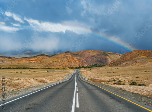 Long winding road leads through dry Altai mountains under dramatic cloudy sky with a vibrant rainbow