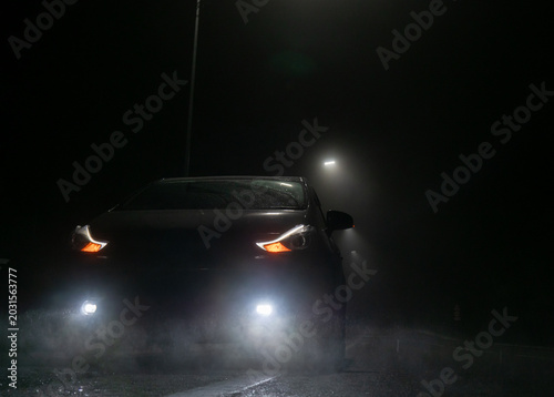 A dark car sits parked on a wet road at night. Its bright headlights, daytime running lights, and fog lights cut through the heavy mist.