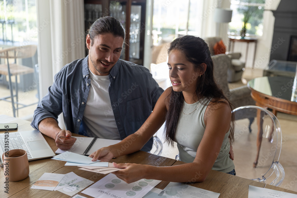 Fototapeta premium Couple sitting at wooden dining table, reviewing printed charts and envelopes with laptop