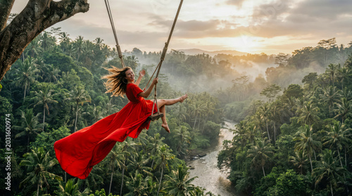 Woman in flowing red dress swinging high over lush green tropical valley