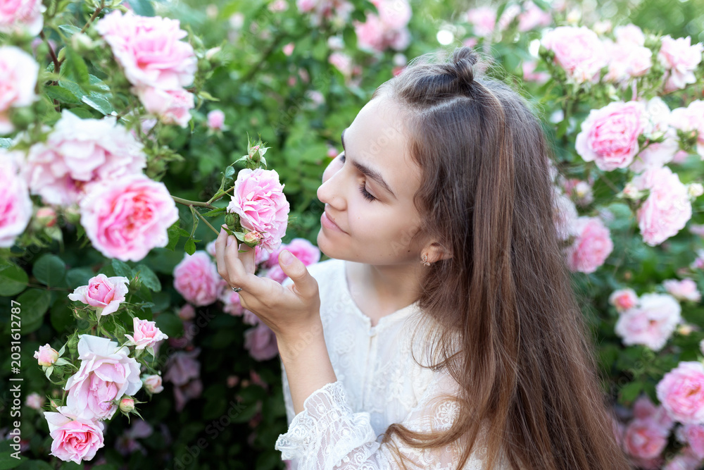 Naklejka premium Beautiful girl smelling a pink rose while in a blooming garden of flowers. Romantic outdoor portrait with long hair, summer atmosphere. Charming lifestyle scene with nature, beauty, and tenderness.