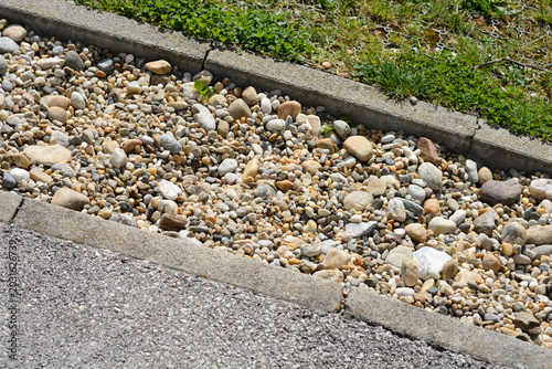 Fotografi A high angle view captures a shallow drainage ditch filled with mixed gravel and river stones situated between a gray asphalt surface and a green grass border