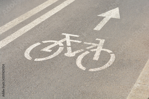 A high angle view captures a white painted stencil of a bicycle and an arrow indicating direction on a gray asphalt bike path next to solid white lines.