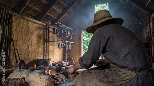 Schilderij op canvas Traditional chef preparing food on an open fire in a reconstructed ancient settlement, depicting historical cooking