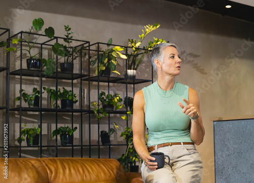 Mature woman sitting on sofa armrest in lounge holding mug, gesturing by plant shelving, copy space