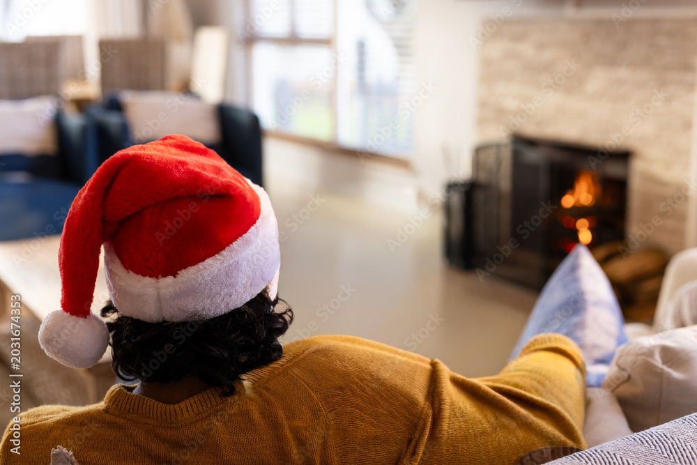 Fototapeta premium Man sitting on light-colored sofa wearing Santa hat and mustard-yellow sweater by fireplace
