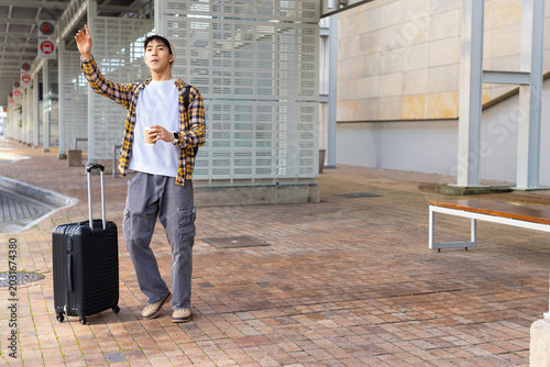 Asian man standing on transit platform with black suitcase and coffee, wearing yellow plaid
