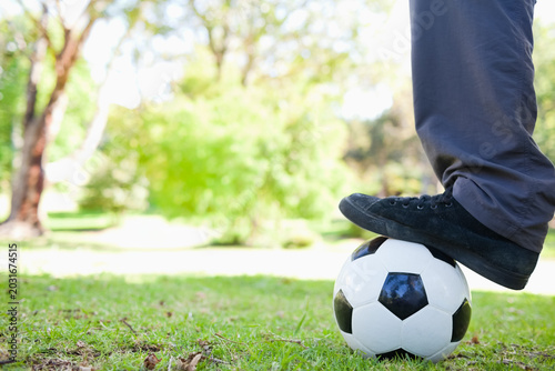 Black sneaker and dark gray trousers are pressing soccer ball on sunlit grassy lawn, copy space
