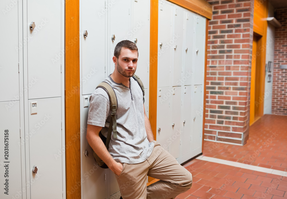 Obraz premium Male student leaning against gray lockers in school corridor, carrying olive backpack, copy space