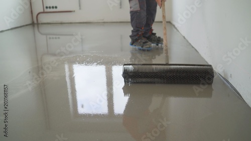 Construction worker applying self leveling screed with spiked roller on concrete floor