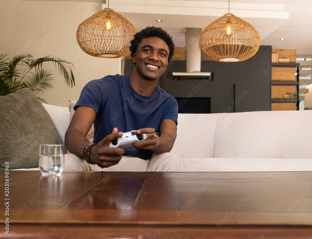 Fototapeta premium African American man sitting on sofa in modern living room holding white controller smiling