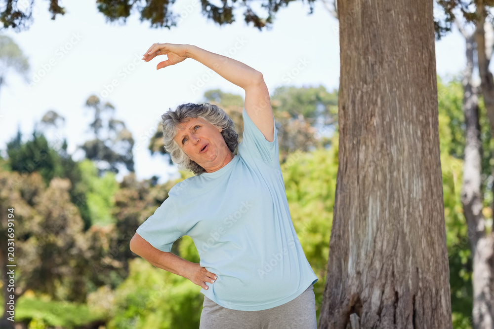 Obraz premium Senior woman doing side stretch beside tree trunk in park wearing light blue shirt and sweatpants