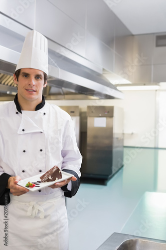 Chef in 20s holding plate with chocolate cake and strawberry, wearing toque in kitchen, copy space