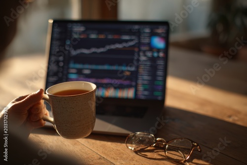 Remote worker sipping tea while analyzing stock market charts on laptop in soft natural light. Home trading, freelance finance, calm workspace. Crypto trading. only hand visible. no face.