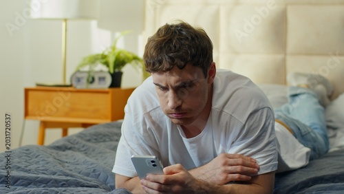 Unhappy Caucasian man lying in bed using smartphone at home worried confused male checking phone problem guy frowning mobile banking fraud spam crash app social media error lost connection breakdown