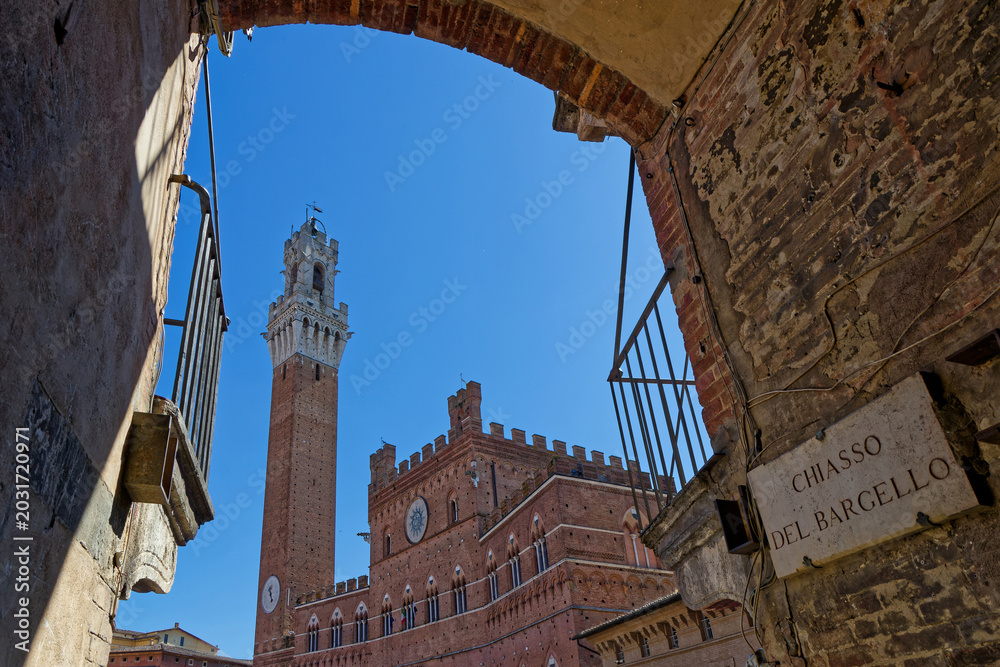 Fototapeta premium SIENA, ITALY, April 25, 2026 : Palazzo Pubblico with its tall Torre del Mangia seen through the frame of an old street arch surrounding Piazza del Campo