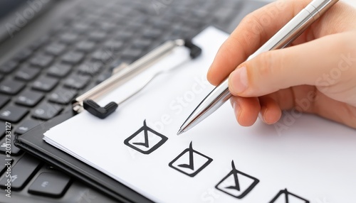 A hand marks checkboxes with a pen on a clipboard placed over a laptop keyboard, symbolizing task completion or survey participation.