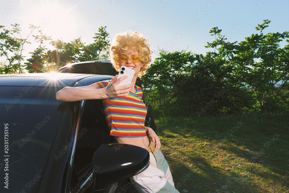 Fototapeta premium Smiling woman in a striped top leans on a car, taking a selfie with a smartphone. Outdoor scene with bright sun, retro film color, and casual fashion mood, under warm golden light and a breezy