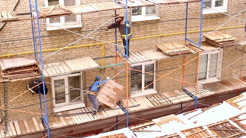 Construction workers moving wooden planks on scaffolding outside a brick apartment building during winter season