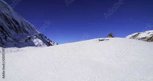 A trekker in a red jacket walks along the snowy shore of stunning Tilicho Lake, framed by towering Himalayan peaks and a brilliant blue sky.