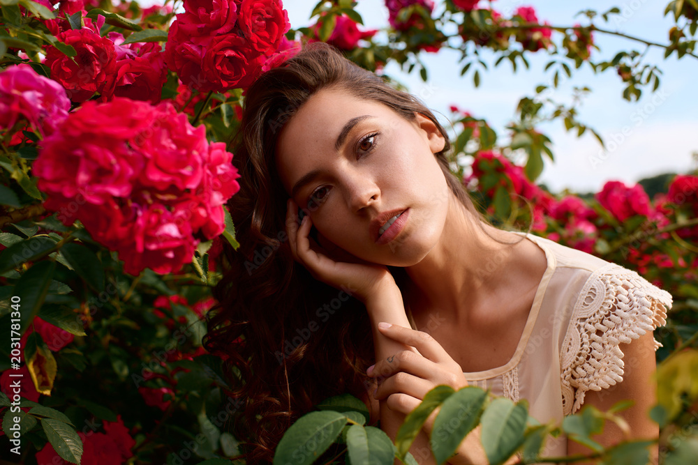 Naklejka premium Enchanting young woman embraces the beauty of nature amidst blooming roses in a sunny park