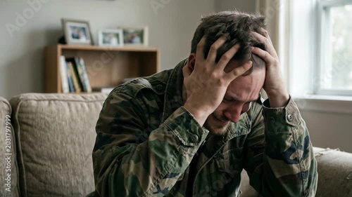 Crying soldier suffering from PTSD in camouflage jacket. Distressed army veteran holding head and weeping on couch. Mental health grief concept