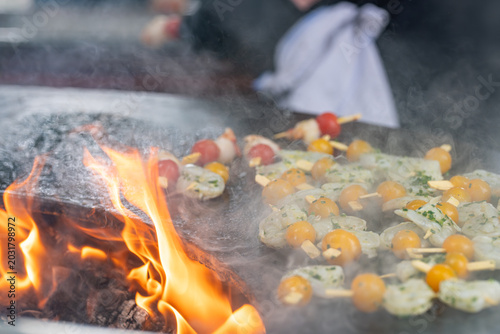 Brochettes de Saint-Jacques et tomates en cuisson sur brasero avec flammes et fumée lors d’un barbecue en extérieur.