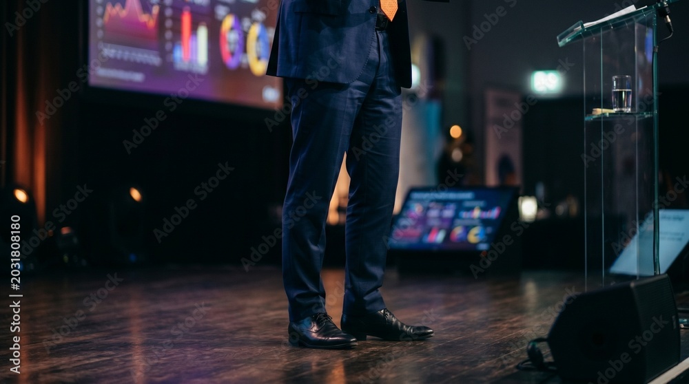 Fototapeta premium Professional businessman standing on stage at a corporate conference giving a presentation with data charts on screen, leadership and high-stakes business communication concept