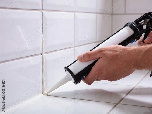 A close-up view of a person applying white sealant from a caulking gun onto a tiled bathroom wall corner.