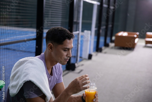 Sitting Indian man holding orange-juice cup on bench by blue court, white towel, wearing purple tee
