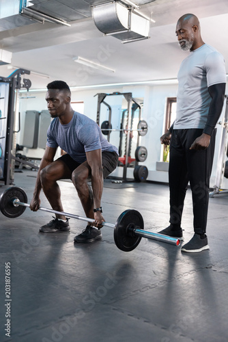 African American men deadlifting with bar and watch while senior trainer coaching, copy space
