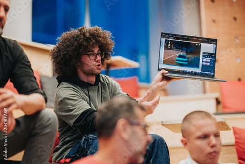 Young man holds a laptop showing a car game while speaking to a small group. The informal tech workshop scene captures collaborative discussion and a product demo.