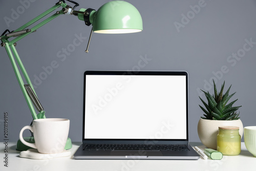 Stylish workplace. Laptop with blank screen, lamp, cup and decor on white table against gray background. Space for text