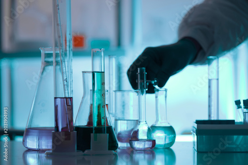 Chemistry laboratory scene with colorful chemical solutions in glass flasks and cylinders, scientist in gloves writing results, analytical experiment workspace.