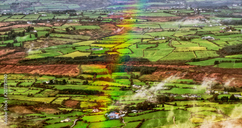 A striking rainbow descends over the vibrant patchwork of green fields and scattered white farmhouses in rural Ireland, observed from above on a clear day.