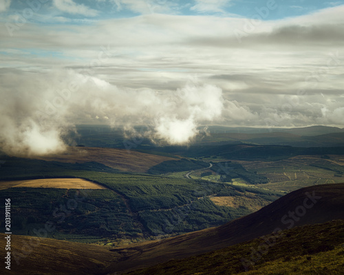 A wide landscape view of rolling green hills and forested areas under a cloudy sky.