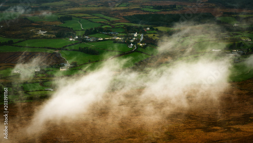 High-angle view of low clouds and mist drifting across a rural landscape of green agricultural fields and small houses.