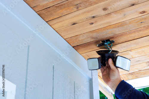 Handy man holds outdoor security lights while preparing to install them on wooden ceiling at their home.