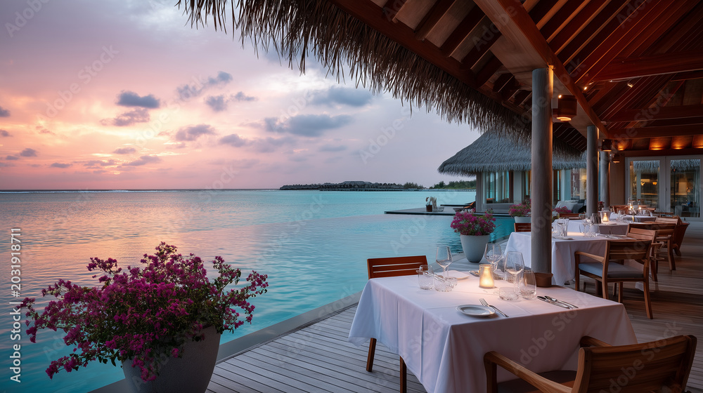 Fototapeta premium Ultra-wide cinematic shot of luxury resort open-air dining pavilion with thatched roof overlooking calm turquoise lagoon at sunset, warm amber and pink evening light washing over w