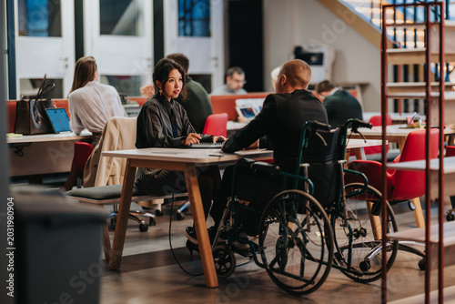 A businesswoman discusses documents with a colleague in a wheelchair at a shared office desk. Colleagues hold a professional meeting in an open-plan workspace.