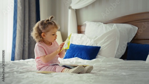 Vászonkép Young girl sits on a cozy bed in pajamas, absorbed in a colorful storybook, turn