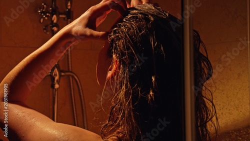 Woman rinsing long wet hair and massaging scalp under warm shower, enjoying relaxing at home haircare routine with water droplets, shampoo foam and soft lighting